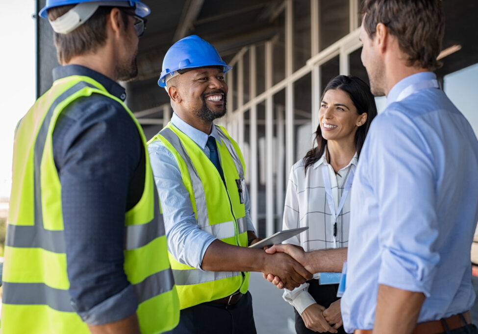 Smiling engineer shaking hands at construction site with happy architect. Handshake between cheerful african construction manager with businessman at bulding site. Team of workers with architects and contractor conclude an agreement with safety uniform.